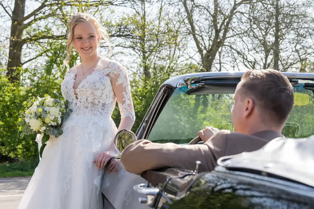 Bruid en bruidegom poseren bij een zwarte cabriolet met het Streekbos op de achtergrond. De bruid draagt een witte kanten jurk en houdt een wit en geel boeket vast. De bruidegom zit in de auto en kijkt naar haar
