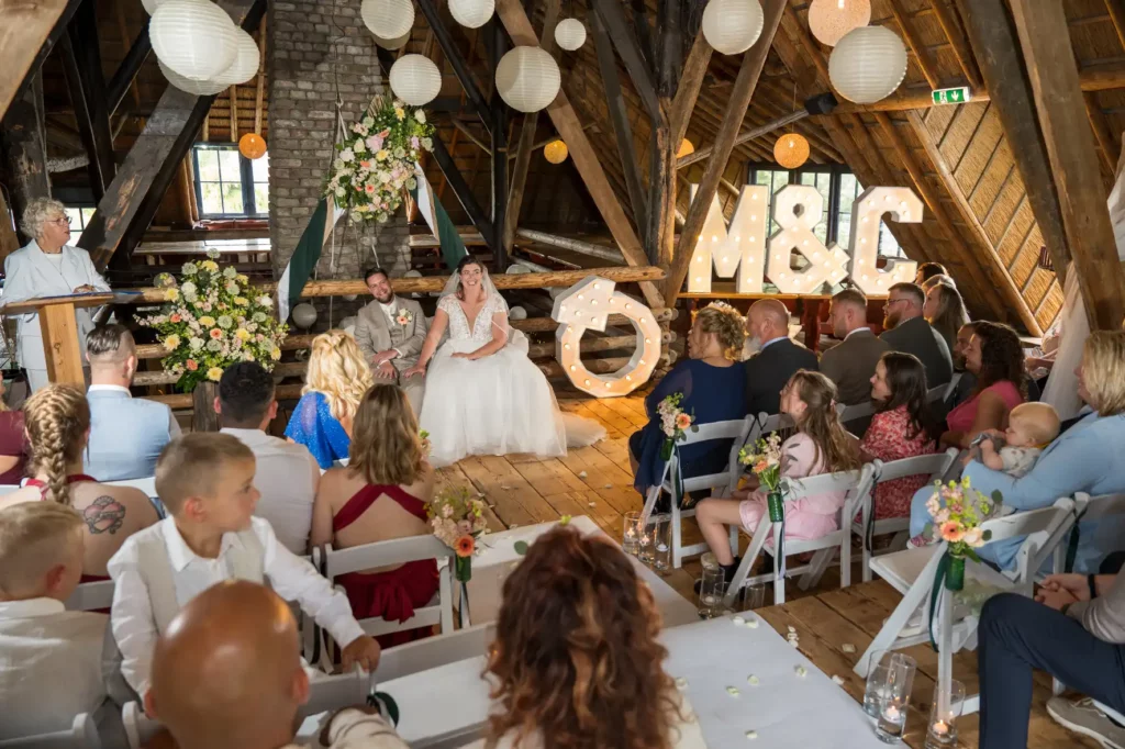 Mick en Charissa zitten samen in de karakteristieke zolder van Robacher's Watermolen tijdens hun huwelijksceremonie. Op de achtergrond staan verlichte letters met 'M&C' en een grote trouwring. Familie en vrienden zijn aanwezig en kijken toe, zittend op witte stoelen. De ruimte is versierd met bloemen en lampionnen