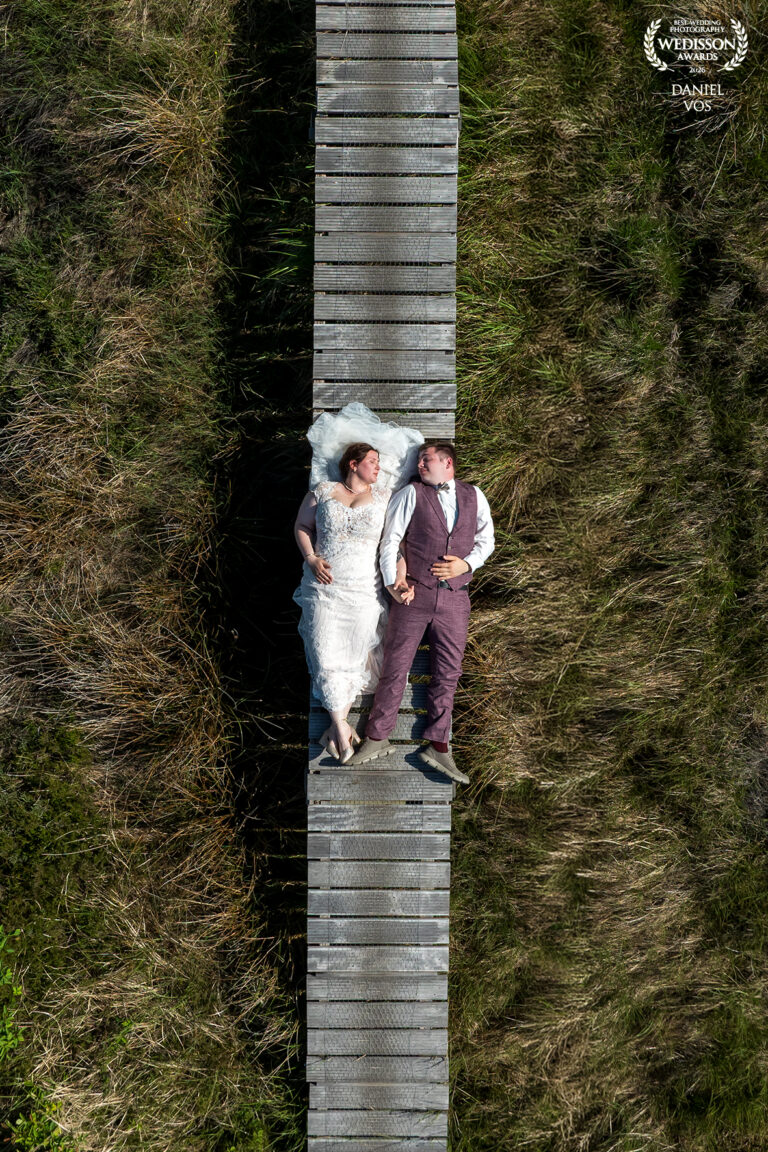 Luchtfoto van bruidspaar Dominique en Jelle liggend op een houten vlonderpad in de natuur van Texel, gefotografeerd door bruidsfotograaf Daniel Vos.