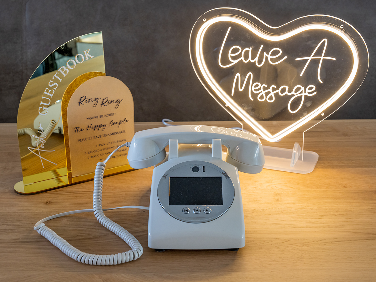 Witte vintage draaitelefoon als audio- en videogastenboek met een verlicht hartvormig neonbord 'Leave A Message' op een houten tafel.